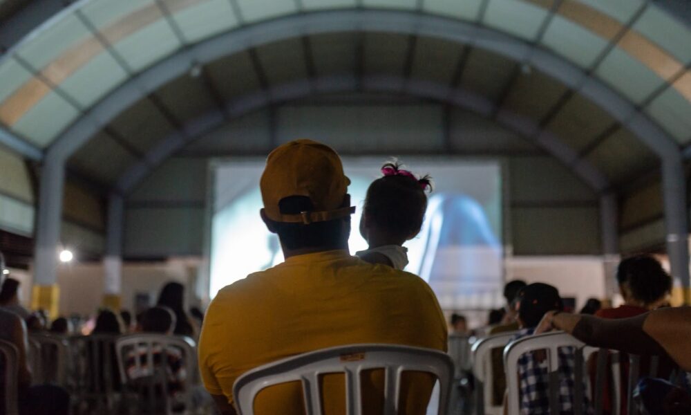Governo de Goiás leva Cine Leitura do Bem a mais quatro municípios - público assistindo a sessão de cinema do projeto. Em primeiro plano, pai com bebê nos braços assistindo ao filme